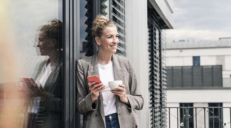 Smiling businesswoman with cell phone and coffee cup standing on roof terrace